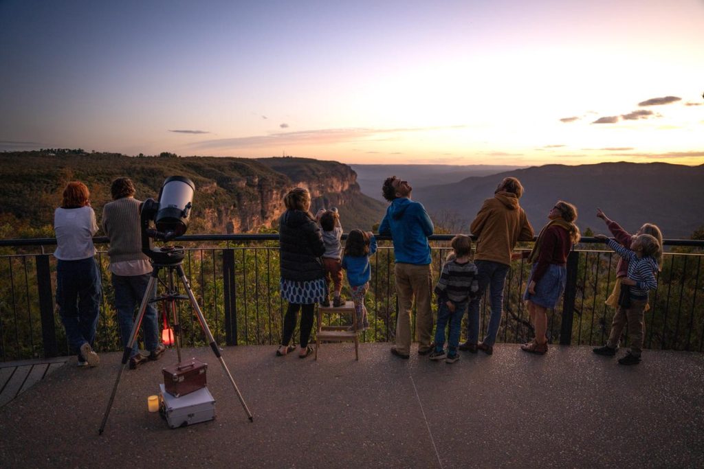 A group of 11 adults and children enjoying the views from Wentworth Falls lookout and looking up and pointing to the sky